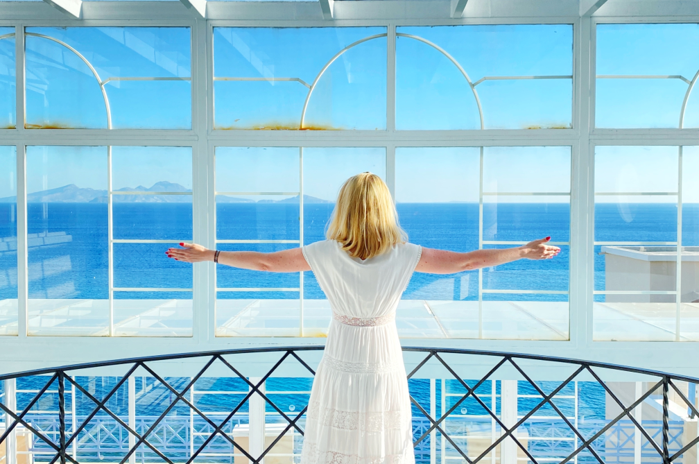 Woman stood on balcony with arms apart looking towards sea through large floor to ceiling windows.